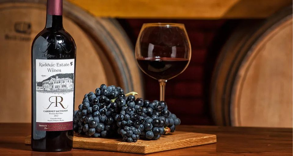 A bottle of the estate Cabernet Sauvignon, a filled glass and a bunch of grapes before the cellar barrels