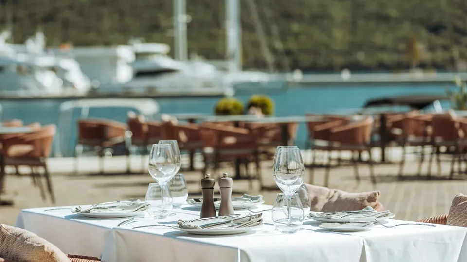 A laid table on the marina terrace, glasses and linen in the foreground, yachts beyond