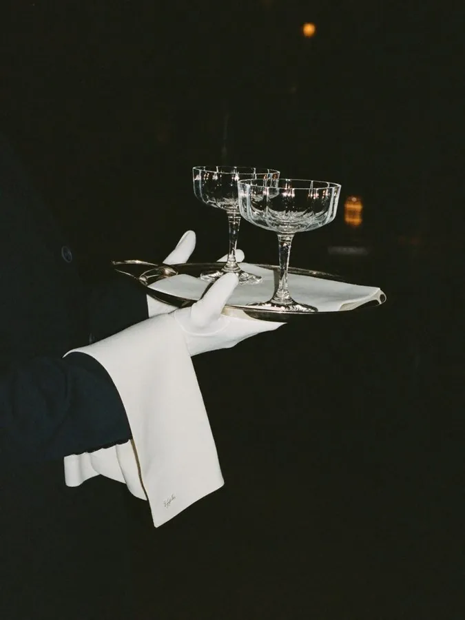 A gloved hand carrying a silver tray with two coupe glasses